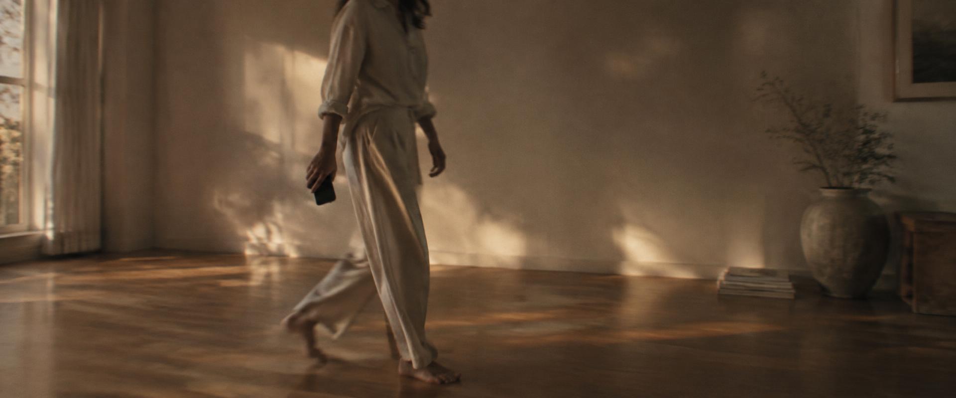 A woman walking barefoot across a sunlit wooden floor with her phone in her hand